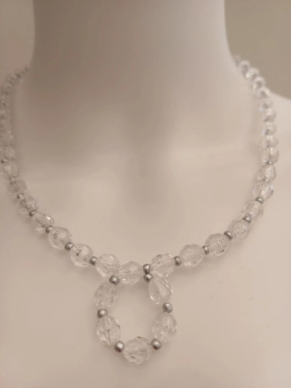 Close-up of a beaded necklace on a mannequin. The necklace features clear, faceted beads with small silver spacers, forming a looped pendant.