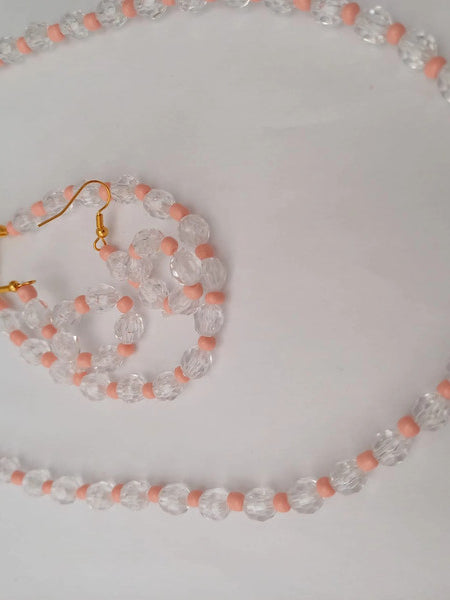 A necklace and earring set made of clear faceted beads and small pink beads, with gold-colored hooks and chain clasps, on a white background.