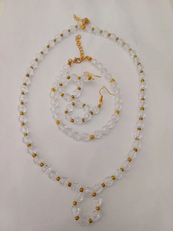 A clear beaded jewelry set on a white surface, featuring a necklace, bracelet, and earrings. The beads are transparent with small gold accents, and the necklace has a gold chain clasp.