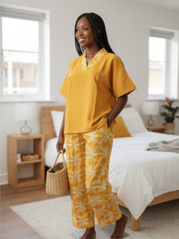 A woman stands in a bedroom wearing a yellow top and patterned yellow pants. She holds a woven basket and smiles. The room has a neatly made bed, two windows, and a small side table with a lamp.