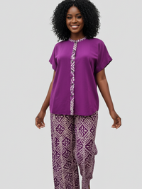 A woman with curly hair smiles, wearing a purple top with a patterned strip and matching purple patterned pants, against a gray background.