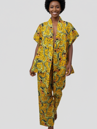 A person with short curly hair wearing a vibrant yellow outfit with intricate green and black swirls and patterns, walking barefoot.