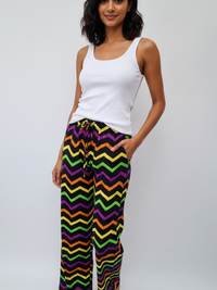 A person stands against a white background, wearing a white tank top and colorful zigzag pattern pants in black, yellow, purple, and green.