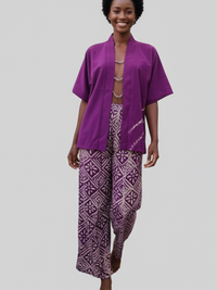 A person with short curly hair is smiling while walking barefoot. They are wearing a purple open-front top and patterned purple and white wide-leg pants against a plain background.