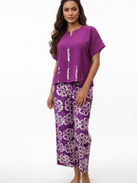 A woman with long, wavy hair is wearing a purple top with white geometric patterns and matching purple pants featuring a white circular design. She stands barefoot against a white background.
