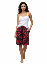 A woman with long curly hair is wearing a white tank top and floral-patterned shorts with red and pink designs, standing barefoot.