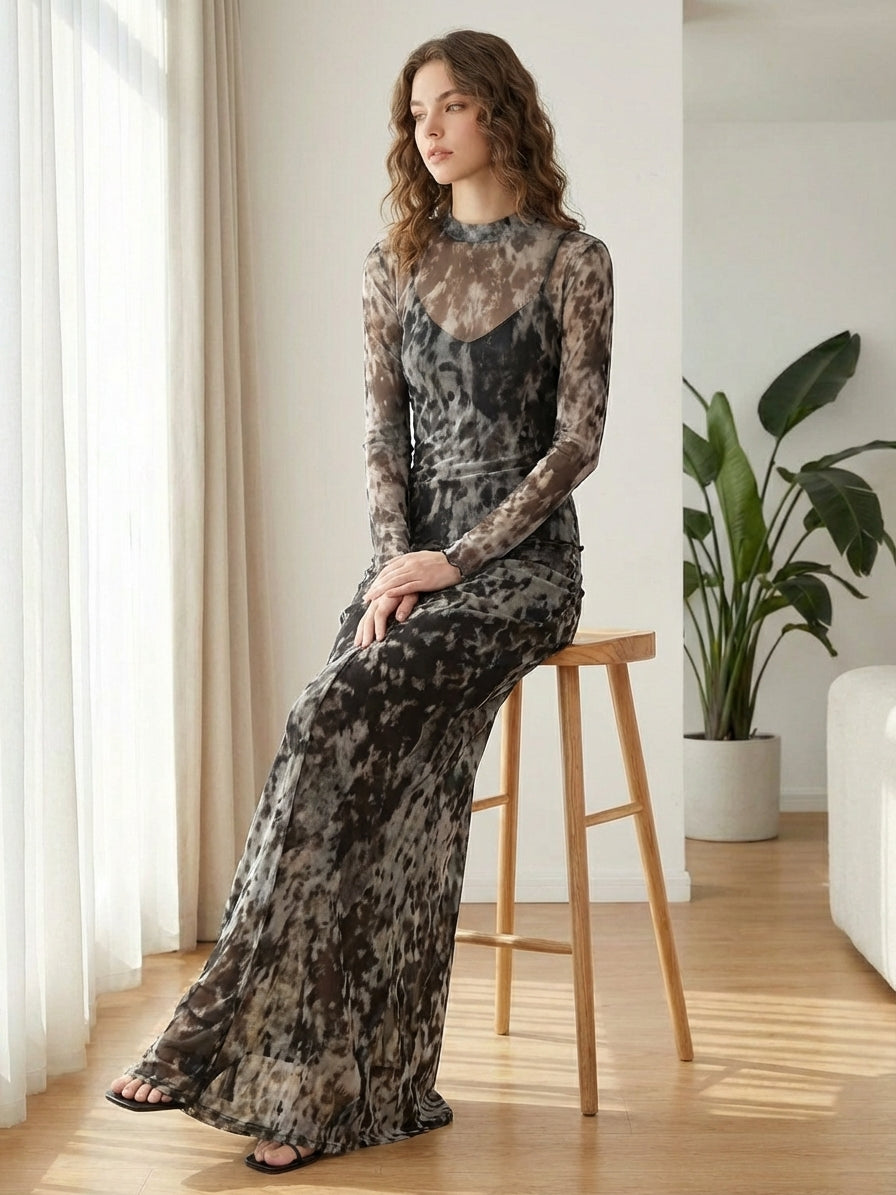 A woman with wavy hair sits on a wooden stool, wearing a long, sheer, patterned dress in shades of gray and black. She is in a modern room with light wooden floors, a large plant, and natural light coming through sheer curtains.