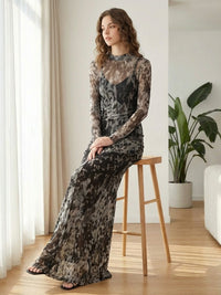 A woman with wavy hair sits on a wooden stool, wearing a long, sheer, patterned dress in shades of gray and black. She is in a modern room with light wooden floors, a large plant, and natural light coming through sheer curtains.