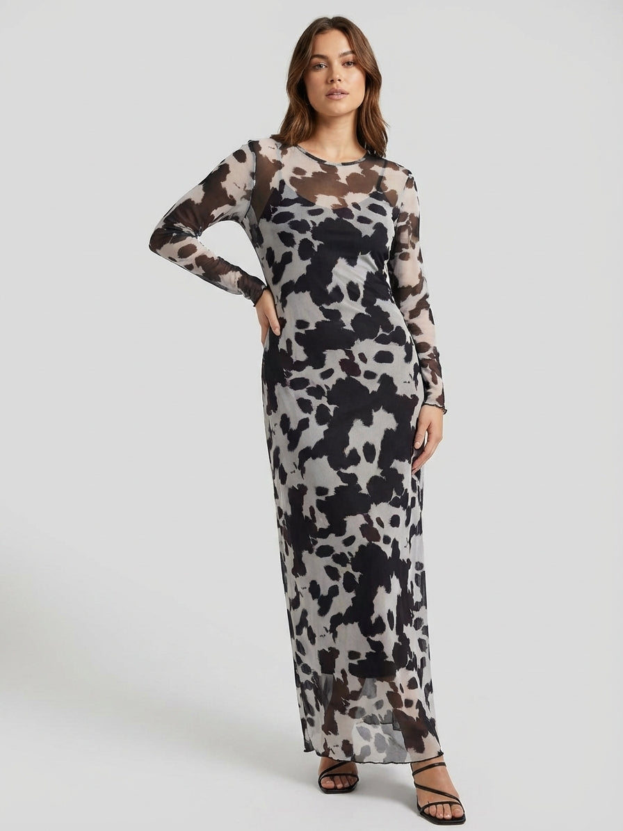 A woman wearing a long-sleeve, black and white abstract patterned dress stands against a plain gray background. She is wearing black strappy sandals.