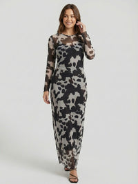 A woman in a long-sleeve, ankle-length dress with a black and gray abstract pattern stands against a plain background. She has brown hair and wears black strappy sandals.