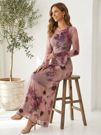 A woman sits on a wooden stool wearing a long-sleeve, tie-dye dress in shades of pink and purple. Her hair is styled in loose waves. An olive tree in a large pot is nearby, against a white wall.