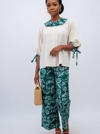 A woman wearing a beige blouse with a green patterned collar and matching wide-leg pants. She holds a woven handbag and wears sandals.