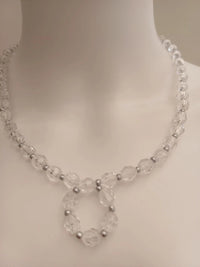 A clear beaded necklace with silver spacer beads, featuring a circular pendant design, displayed on a white mannequin bust.