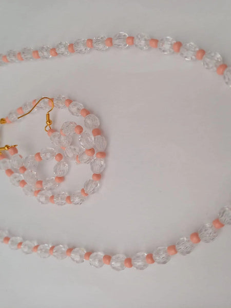 A jewelry set featuring a clear and peach bead necklace with matching earrings. The beads are round and faceted, connected by small peach beads. The necklace has a gold clasp and chain extender.
