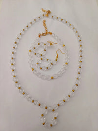 A clear beaded jewelry set on a white surface, featuring a necklace, bracelet, and earrings. The beads are transparent with small gold accents, and the necklace has a gold chain clasp.