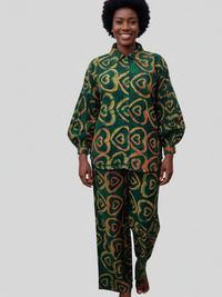 A person with short curly hair is smiling, wearing a green outfit with a yellow and red heart pattern. The background is plain gray.