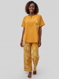 A person smiling, wearing a mustard yellow top with a patterned pocket and matching wide-leg pants featuring a leaf design. The background is plain light gray.