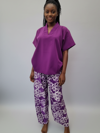 A woman with braided hair stands barefoot against a plain background, wearing a purple top and patterned purple pants with white designs.