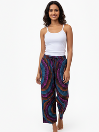 A woman with shoulder-length hair smiles while walking barefoot. She wears a white tank top and colorful, patterned pants with blue, purple, and orange swirls. The background is plain white.