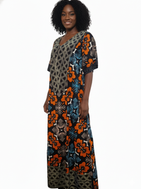 A woman with curly hair smiles, wearing the African Print Maxi Dress – Vibrant Floral Pattern, featuring bold orange, blue, and black floral and geometric designs, standing against a white background.