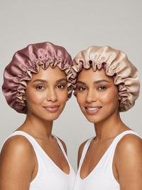 Two people wearing satin bonnets, one in rose pink and the other in beige. They are smiling and dressed in white tank tops against a neutral background.