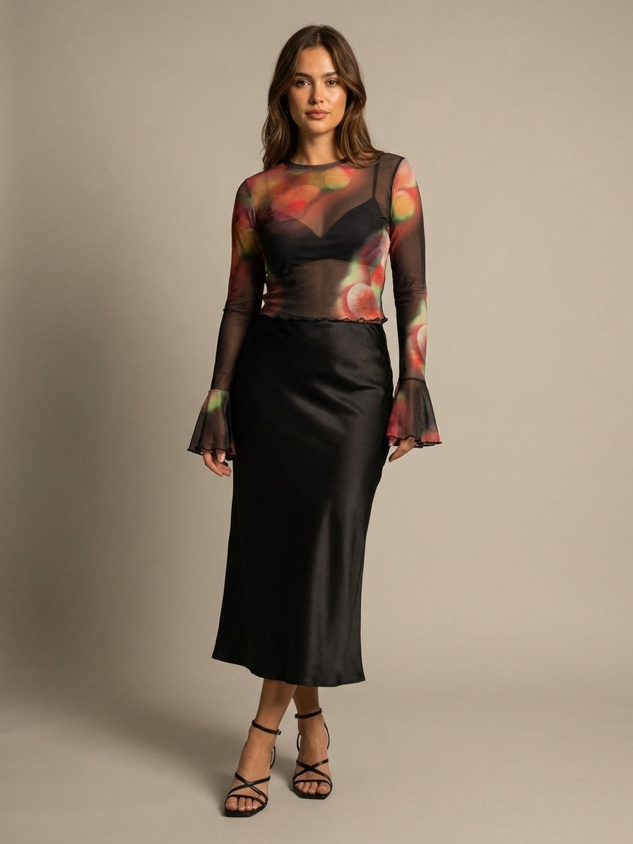 A woman stands against a neutral background wearing a sheer, colorful top with bell sleeves and a black satin skirt. She has long brown hair and is wearing black strappy sandals.