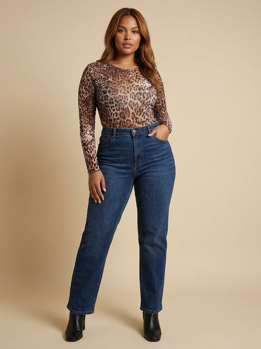 A woman stands against a beige background wearing a leopard print long-sleeve top, blue jeans, and black ankle boots.