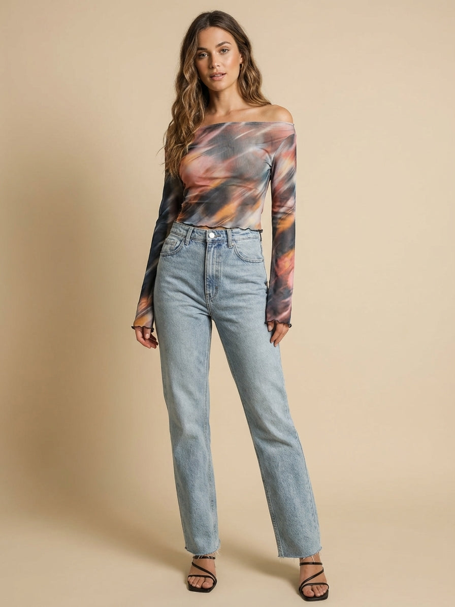 A woman stands against a beige background, wearing a long-sleeve, off-the-shoulder top with a colorful, swirling pattern and light blue jeans. She has long, wavy hair and is wearing black strappy sandals.