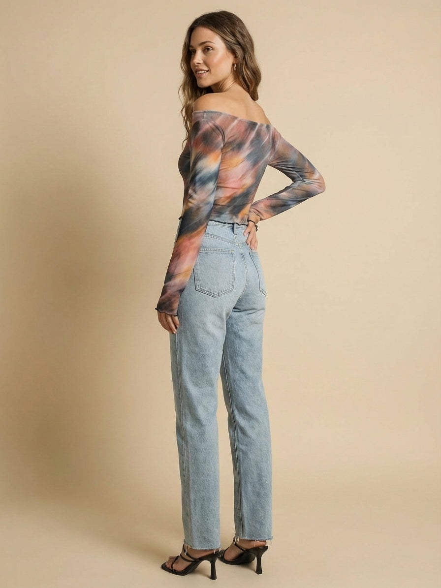 A woman with long hair wearing a colorful off-shoulder top, light blue jeans, and black heeled sandals stands against a beige background, looking over her shoulder.