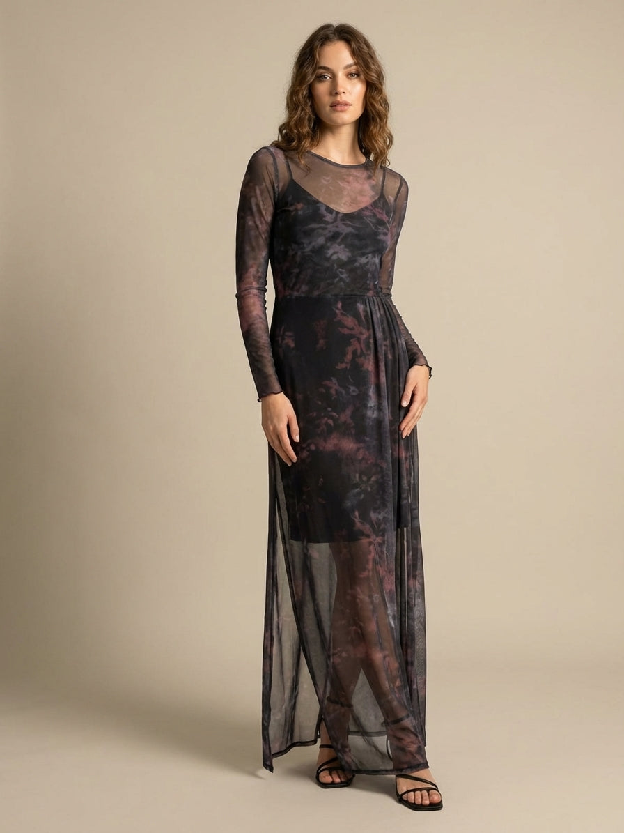 A woman with wavy hair wearing a long, sheer, dark tie-dye dress with long sleeves, standing against a beige background. She is wearing black sandals.