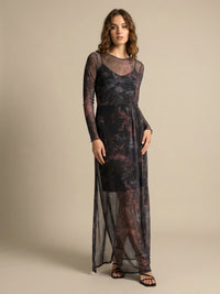 A woman with wavy hair wearing a long, sheer, dark tie-dye dress with long sleeves, standing against a beige background. She is wearing black sandals.