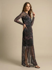 A woman in a long-sleeved, sheer black dress with a purple and pink abstract pattern stands against a beige background. She has wavy brown hair and wears black strappy heels. Her right hand rests on her hip, while she gently touches her hair with her left hand.