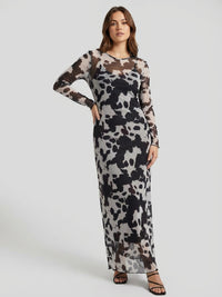 A woman wearing a long-sleeve, black and white abstract patterned dress stands against a plain gray background. She is wearing black strappy sandals.