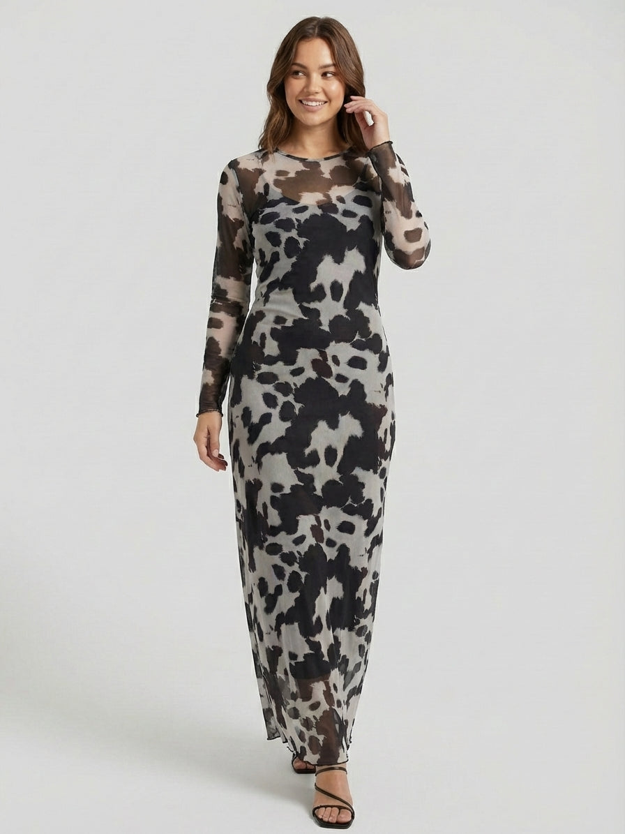 A woman in a long-sleeve, ankle-length dress with a black and gray abstract pattern stands against a plain background. She has brown hair and wears black strappy sandals.