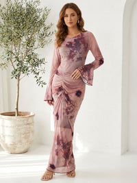 A woman in a long-sleeve, tie-dye pink and purple dress with flared cuffs stands next to a potted olive tree on a white background.