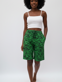 A woman with curly hair wearing a white tank top and green zebra-striped shorts stands barefoot against a white background.